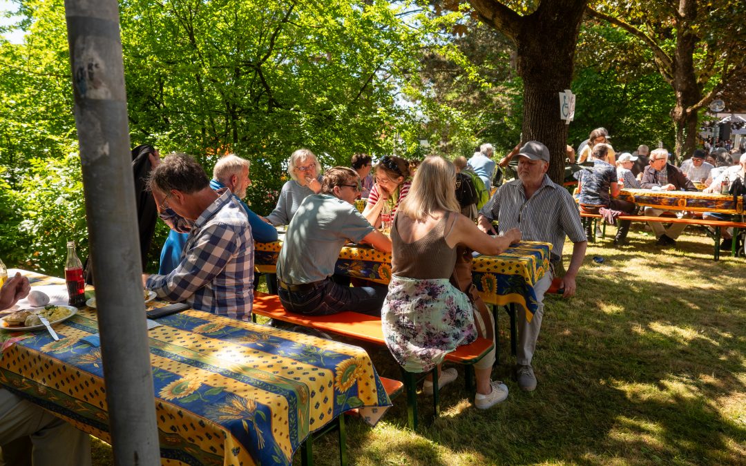 Strahlender Sonnenschein beim Karlshöher Jahresfest. Menschen sitzen auf Biertischen auf einer saftigen Wiese und essen.